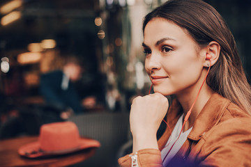 Portrait of woman sitting at restraunt and relaxing. She is holding hand close to chin and thinking. Girl is enjoying the moment. Her hat is lying on table