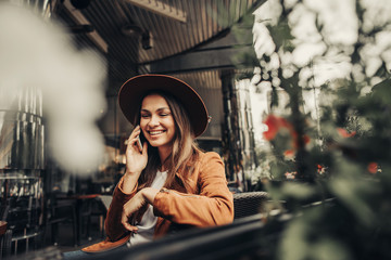 Cheerful woman is sitting on bench and talking on the phone. She is looking down and smiling. Girl wears brown jacket and hat