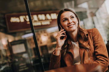 Attractive womn in brown jacket is sitting and talking on the phone. She is looking to the right. Girl is sitting at table in restaurant