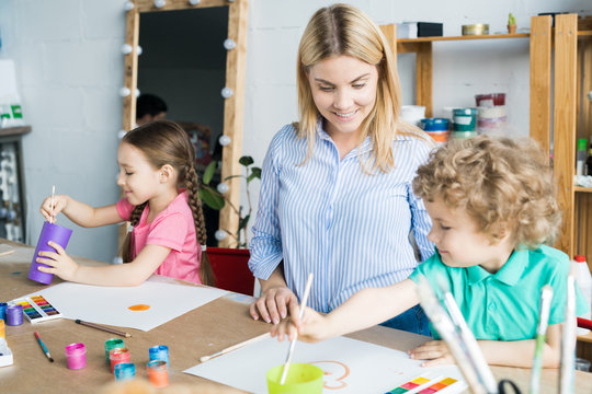 Smiling young teacher in art studio helping children in painting