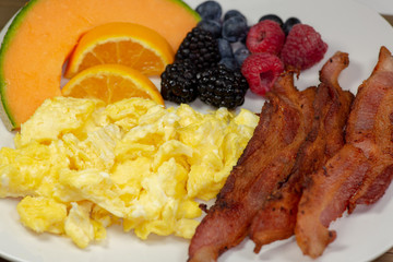Breakfast plate of bacon, cantaloup slice, orange slices, blackberries,  raspberries and blueberries on the kitchen table waiting to be eaten