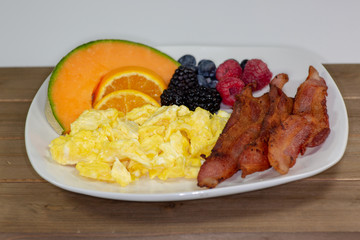 Breakfast plate of bacon, cantaloup slice, orange slices, blackberries,  raspberries and blueberries on the kitchen table waiting to be eaten
