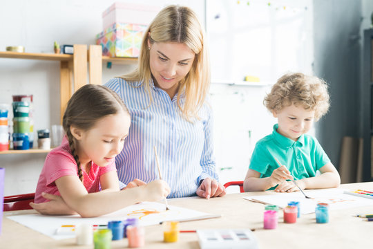 Two kids, boy and girl, painting together with their art teacher at the table at art class