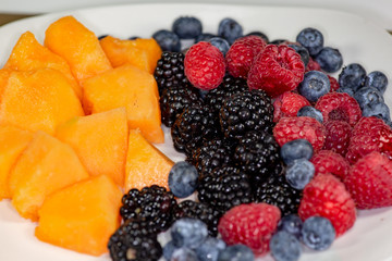 A plate of cantaloup, blueberries, raspberries, blackberries on a white plate sitting on the kitchen table waiting to be eaten
