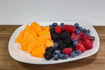 A plate of cantaloup, blueberries, raspberries, blackberries on a white plate sitting on the kitchen table waiting to be eaten