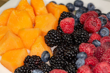 A plate of cantaloup, blueberries, raspberries, blackberries on a white plate sitting on the kitchen table waiting to be eaten