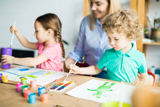 Little boy with paintbrush drawing his picture with paints together with his sister and mother helping to them