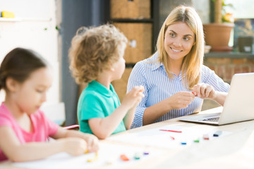 Smiling teacher sitting at the table with laptop and sculpting from play clay together with pupils