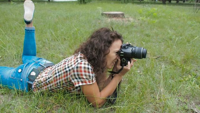 A female photographer is working in the park. A woman with a camera lies on the grass.