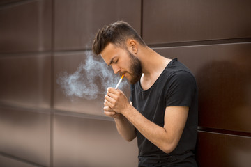 A young handsome white bearded man in black t-shirt smokes a cigarette in the street in the spring....