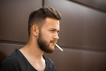 A young handsome white bearded man in black t-shirt smokes a cigarette in the street in the spring. Close up.