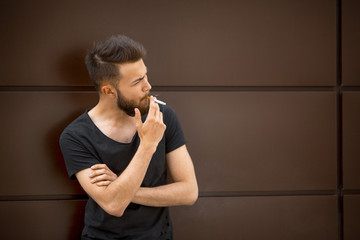 A young handsome white bearded man in black t-shirt smokes a cigarette in the street in the spring. Close up.