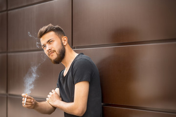A young handsome white bearded man in black t-shirt smokes a cigarette in the street in the spring. Close up.