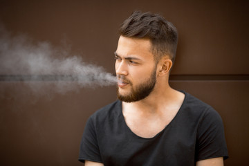 A young handsome white bearded man in black t-shirt smokes a cigarette in the street in the spring. Close up.