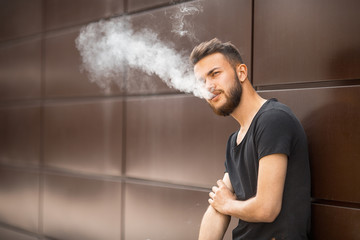 A young handsome white bearded man in black t-shirt smokes a cigarette in the street in the spring. Close up.
