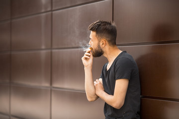 A young handsome white bearded man in black t-shirt smokes a cigarette in the street in the spring. Close up.