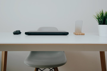 Office place, office with a white table, chair. Decorated white desk with black laptop, computer mouse, mobile phone and flower in a pot. Workplace concept.