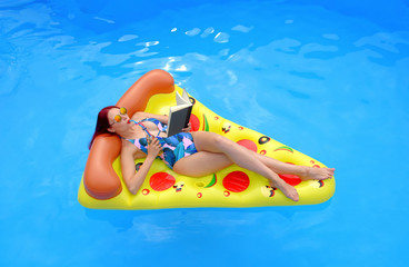 A young woman wearing a bathing suit lies relaxed on an  inflatable swim bed. She takes time out to read a book while she enjoys the pool atmosphere.