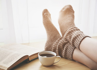 Woman resting keeping legs in warm socks on table with morning coffee and reading book