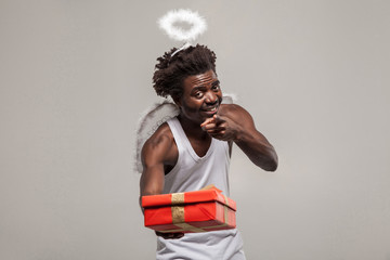 pointing finger sharing Love and gift. dark skinned bearded African angel funny man with white shirt, nimbus and winks atanding and holding gift box. its for you. Indoor, isolated on gray background - obrazy, fototapety, plakaty