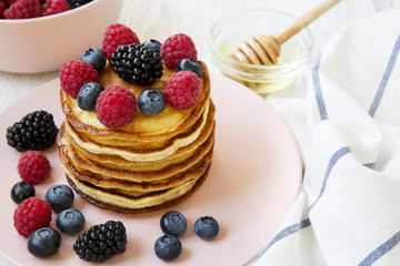 Pancakes with berries and honey on a pink plate over white wooden background, side view. Close-up.