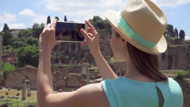 Woman in hat near ancient ruins Forum Romanum taking photo on mobile phone in slow motion. Happy female tourist taking picture of Roman forum in center of Rome, Italy. Student travel through Europe