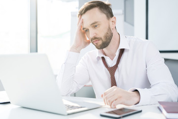 Portrait of tired handsome businessman wearing shirt and tie using laptop while working at desk in office
