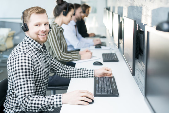 Portrait of smiling young man wearing headset looking at camera while working with group of help desk operators sitting in row, copy space