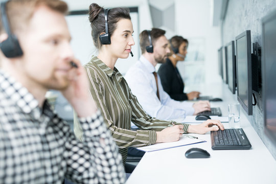 Side view portrait of several help desk operators wearing headsets sitting in row looking at computer screens and talking to clients