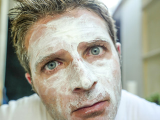 young funny attractive man at home bathroom looking at toilet mirror applying facemask in his face finding himself weird and ugly with the white mask