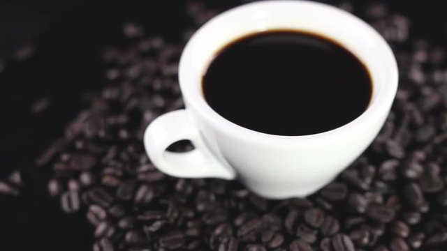 Black coffee in white cup with coffee beans rotating on dark surface.