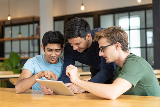 Positive developers discussing their startup during break. Three cheerful young men sharing tablet, modern office interior in background. Teamwork concept