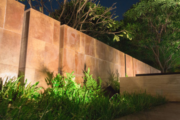 Soft shadows casting onto a landscaped wall with nice mood lighting in the evening near the swimming pool.  Nice design and layout with green grass and plants near lush trees.