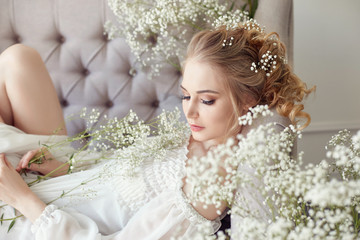 Girl white light dress and curly hair, portrait of woman with flowers at home near the window, purity and innocence. Curly blonde romantic look, beautiful eyes. White wildflowers in hands