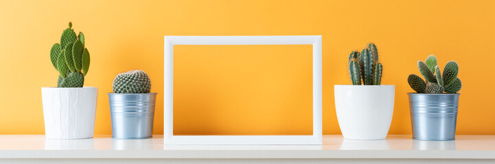 Collection of various cactus plants in different pots. Potted cactus house plants on white shelf against pastel mustard colored wall and picture frame mock up banner.