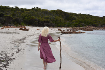 full length portrait of blonde girl wearing purple dress, walking along a beach.