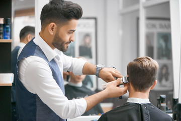 Barber doing new haircut for young client sitting in front of mirror. Wearing white casual shirt,...
