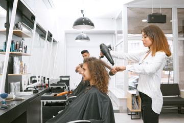 Female hairstylist drying curly girl's hair using big plastic brush and electric dryer. Wearing white casual shirt and black high waist trousers. Client sitting near mirror covered with black cape.