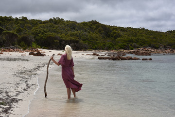 full length portrait of blonde girl wearing purple dress, walking along a beach.