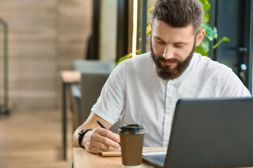 Bearded hipster writing notes while working with laptop, drinking coffee. Wearing white casual shirt, leather bracelet, having dark brown hair. Stylish interior of modern restaurant on background.
