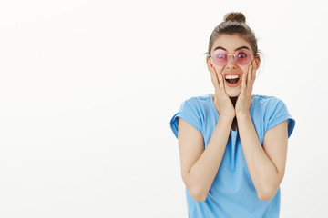 Naklejka premium Studio shot of emotive excited beautiful and funny woman in trendy sunglasses and casual summer t-shirt, staring at camera and touching face with palms, being surprised and over-emotive