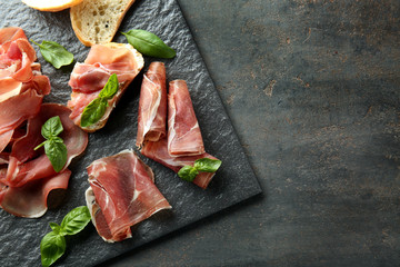 Slate board with tasty prosciutto slices and basil on table