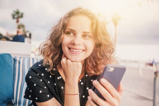 Young beautiful happy smiling woman with a smartphone in hands sitting in a street cafe, close-up portrait, shot of a head