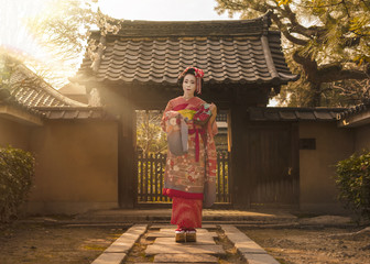 Maiko in a kimono posing on a stone path in front of the gate of a traditional Japanese house...