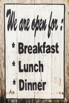 Wooden textured board with painted handwritten black text "We are open for Breakfast, Lunch, Dinner"