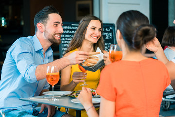 Cheerful young best friends toasting with a refreshing delicious summer drink while sitting together at table at a trendy restaurant outdoors