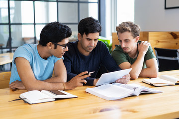 Focused students using tablet and discussing information. Young men studying and sitting at desk in classroom or library. Education and technology concept.
