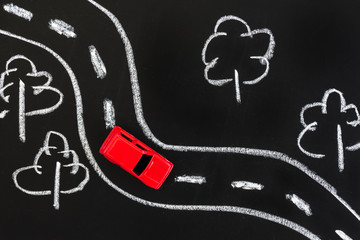 Red toy car on a blackboard with chalk drawn road and trees.