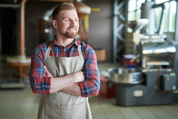 Waist up portrait of cheerful bearded man wearing apron posing standing confidently with arms crossed against roasting machines in artisan coffee roastery, copy space