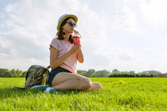 Young girl in sunglasses and hat drinks summer berry drink with ice sitting on green grass. Copy space.